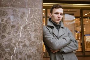 A young man stands outside a shop on the street in a gray coat leaning. photo