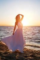 hermosa mujer posando en el playa a puesta de sol en un blanco vestir foto