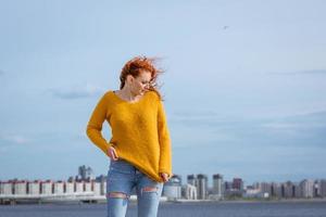 Red-haired woman poses against river in city with buildings in background. photo