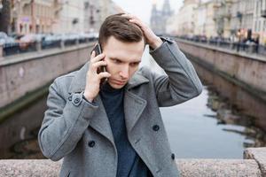 Handsome young man using mobile phone, wearing an elegant gray coat, stands photo