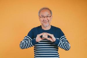 Senior male holding wooden house model property owner after retirement on yellow background. photo