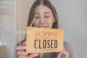 Smiling asian young owner retail, coffee shop woman turning sign board to closed after finished work, job and protection to pandemic of coronavirus. Close store due to lockdown, quarantine of covid. photo