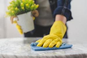 limpieza higiene, mano de mucama, camarera mujer vistiendo amarillo protector guantes mientras limpieza , tomar fuera maceta desde mesa y utilizando azul trapo limpiando a polvo. limpieza interna limpiar arriba, limpiador. foto