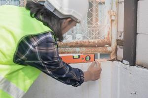 mano de joven asiático contratista, constructor hombre utilizar agua nivel metro medición el muro, inspeccionar el reconstruido a construcción y renovación hogar, cheque defecto a balcón. ingeniero trabajador concepto. foto