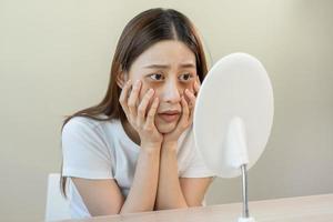 Bored, insomnia asian young woman, girl looking at mirror hand touching under eyes with problem of black circles or panda puffy, swollen and wrinkle on face. Sleepless, sleepy healthcare person. photo