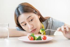 anorexia, infeliz hermosa asiático joven mujer, niña en haciendo dieta, participación tenedor a brócoli en ensalada lámina, disgusto o cansado con comer Fresco vegetales. nutrición de bueno saludable, aburrido con alimento. foto