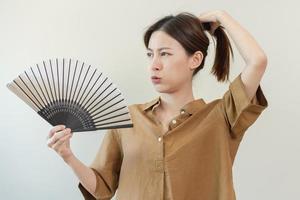 Suffering summer heat stroke, hot weather, tired asian young woman, girl sweaty and thirsty, refreshing with hand in blowing, wave fan to ventilation when temperature high at home, house on background photo