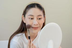 Happy routine beauty concept, pretty asian young woman, girl make up face by applying powder foundation by brush around face, looking at the mirror at home. Female look with natural fashion style. photo