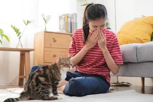 Asian young woman, girl hand in sneezing from fur allergy while playing with her lovely cat, pet on carpet in living room at home, apartment. Health care rhinitis diseases allergic to animal hair. photo