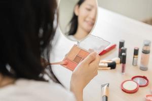 Happy beauty, beautiful asian young woman, girl hand applying brush blush on color powder case when makeup on face in routine, looking in mirror at home, look with natural fashion style, top view. photo