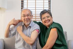 coronavirus vacunación, contento asiático anciano, Envejecido familia sonrisa fuerte juntos, demostración vendaje en brazo con proteger de covid-19 después inyección de vacuna, sentado en sofá en vivo habitación a hogar.