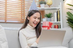 Celebrate on day of happy birthday season, pretty asian young woman using laptop computer, greeting on video call during social distance, holding gift box, happy to get present on couch, sofa at home. photo