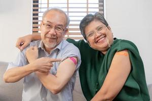 Coronavirus Vaccination, happy asian elderly, aged family smile strong together, showing bandage on arm with protect of covid-19 after injection of vaccine, sitting on couch in living room at home. photo