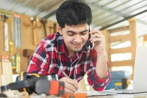 Owner entrepreneur startup small shop business. Carpenter asian young man using smartphone, mobile talk with client about order, use laptop computer for marketing online. Factory Industry of wood. photo