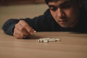 Stressed, sad asian young man, male holding medicine, lying on table. Close up hand of abuse overdose pills and addict. Sick pain of health treatment, unhappy people. Suicide depressed or despair. photo