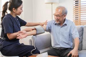 Pressure measurement of hypertension in elderly man sitting on couch, young woman nurse or caregiver care checking, measuring heart, blood by using sphygmomanometer monitor at home after retirement. photo