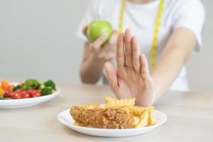 dieta, dieta asiático joven mujer mano en empujar afuera, negar frito pollo, francés frito y escoger verde manzana, vegetales ensalada, comer comida para bueno saludable, salud cuando hambriento. hembra peso pérdida gente. foto