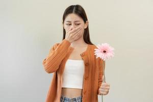 Allergic rhinitis symptom of odor pollen flowering, portrait asian young woman hand in sneeze, blowing runny nose after smell, smelly from bloom flower, holding away pink flora on white background. photo