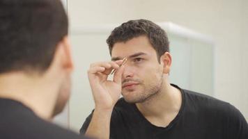 Taking eyebrows with tweezers. Personal care. Young man looking in the mirror and plucking his eyebrows with tweezers. video