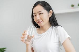 Happy beautiful, asian young woman, girl drinking, sip fresh glass of water for hydration of body, holding transparent glass in her hand, thirsty at home. Health care, healthy lifestyle concept. photo