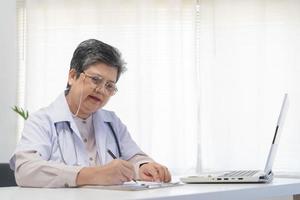 mayor asiático médico, general mujer médico vistiendo lentes y blanco uniforme, maduro hembra con estetoscopio, trabajando en ordenador portátil computadora, sentado en médico oficina. cuidado de la salud y médico. foto