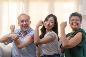 coronavirus vacunación, contento asiático mayor familia con hija sonrisa fuerte juntos, demostración vendaje en brazo proteger de covid-19 después inyección de vacuna, sentado en sofá en vivo habitación a hogar.