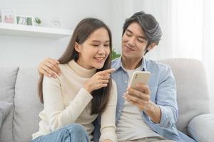 Happy couple love at home,beautiful two asian young spending good time together,bonding to each other and smiling romantic on sofa in living room while man embrace woman using smartphone, mobile phone photo