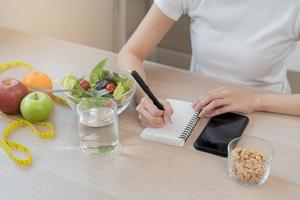 dieta, dieta mano de asiático joven mujer laboral, escribir dieta plan Derecha nutrición con comer Fresco vegetales ensalada es comida para bueno salud no comer basura alimento. nutricionista femenino, peso pérdida de saludable. foto