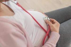 Prenatal, maternity Asian young pregnant woman using stethoscope to listen to her child's heartbeat, making on belly, abdomen. Healthcare of maternal while baby unborn. Wellbeing of motherhood. photo