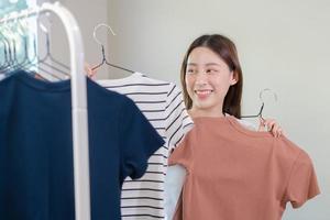 elección de ropa, nada a tener puesto. atractivo asiático joven mujer, niña tratar en aparecer, contento elegir vestido, atuendo en percha en guardarropa en habitación armario a hogar. decidiendo blusa qué a poner en cuales uno foto