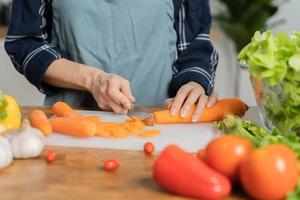 asiático joven mujer, niña o ama de casa utilizando cuchillo, corte zanahorias en junta, en de madera mesa en cocina hogar, preparando ingrediente, receta Fresco vegetales para Cocinando comida. sano comida gente. foto