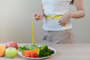 Slim shape asian young woman, girl standing, hand in measure tape around waist, body her have a vegetables on table as white background. Diet session for wellbeing health, eat good food people. photo