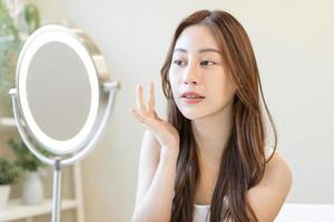 Fresh healthy skin, beautiful smile of asian young woman, girl looking at mirror, applying moisturizer on her face, putting cream treatment before make up cosmetic routine at home. Facial Beauty. photo