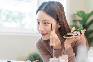 Beauty blogger concept, beautiful and cute asian young woman, girl makeup face by applying brush for eyes on eyebrow, eyeshadow looking at the mirror at home. Female look with natural fashion style. photo