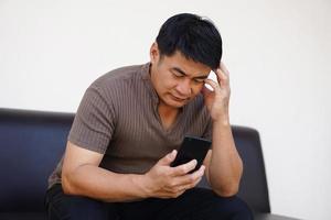 Asian man sits on sofa, hold smartphone, feels headache. Upset, sad. Concept, know bad news from social media online. photo