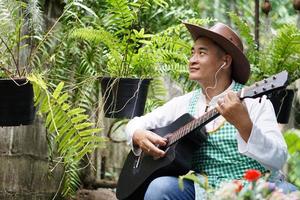Asian man is playing the acoustic guitar in his home back yard garden. Concept, recreation activity, relax time with music and nature. Hobby, pastime. Love song and music. Easy living lifestyle.