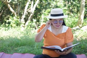 Asian middle- aged woman sits at the park, reads book, holds eyeglasses, has difficulties to see text because of bad vision. Concept , Eyesight problem. suffering eyestrain from reading  book. photo