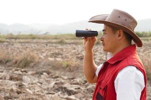 Asian man explorer wears hat, red vest shirt, holds binocular to explore boundary land. Concept,  land property exploration. Long vision distance, zoom for clearing  scene. Observation for something photo