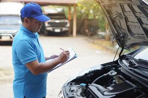 asiático hombre mecánico usa azul gorra y azul camisa, sostiene papel, comprobación y analizando coche motor debajo el capucha. concepto, al aire libre coche inspección servicio. Reclamación para accidente seguro. foto