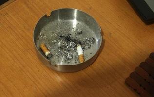 Couple of cigarettes with ashes on metal iron ashtray on top of wooden desk. Isolated photo with landscape background.