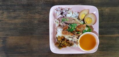 Flat lay or top view stewed pork leg with rice, garlic, slices of boiled egg, pickle, Vegetable and sour sauce in pink dish on dark brown wooden background or table. Thai local and Street food photo