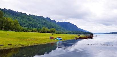 Beautiful of landscape view. Long tail boat parked or floating on the water with green grass, tree, big mountain and cloud sky background at Srinakarin lake, Kanchanaburi, Thailand. Nature wallpaper photo