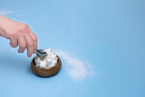 hand with tongs reaches for lump sugar in a wooden bowl on a blue background with copy space photo