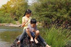 Asian girls and boys take a break from playing in the river to sit on the rocks. Both of them laughed and dodged the water that had been poured over them. Water droplets spread and look fresh. photo