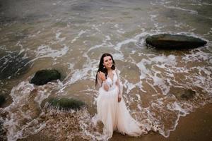 romántico hermosa novia en blanco vestir posando en terraza con mar y montañas en antecedentes foto