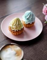 Two delicious, Sweet cream Cakes on a pink plate with yellow and blue cream sit on a wooden table. A cup of cappuccino with wooden Capcakes on the background. Close-up. Copy space photo
