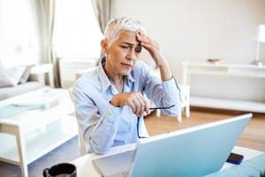 mayor frustrado mujer trabajando desde hogar oficina en frente de computadora portátil, teniendo un dolor de cabeza mujer teniendo un doloroso cara expresión