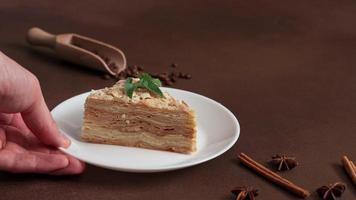 Man's hand Picks up white plate of Layered Cake with cream Napoleon with mint on Brown background. A cinnamon stick, badyan, coffee beans on a brown background. Copy space video