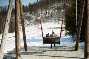 Back view of older brother with younger sister swinging on big wooden swing in forest of mountains. photo