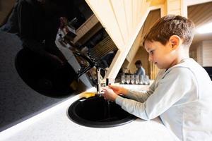 Boy washes hands in the sink at kitchen  in cozy wooden tiny cabin house. Life in countryside.